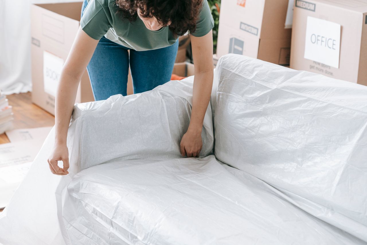 Une femme emballe soigneusement un canapé dans un matériau de protection tout en préparant des cartons dans une pièce lumineuse.