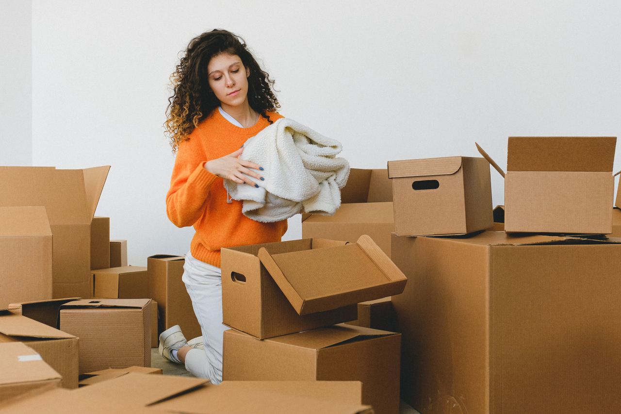 expertise-01 Jeune femme concentrée aux cheveux bouclés, à genoux sur le sol, en train de ranger des vêtements dans des cartons avant de déménager dans un nouvel appartement